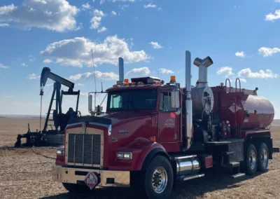 A red Kenworth tanker truck is parked in a vast, dry field under a partly cloudy blue sky. An oil pumpjack is visible in the background